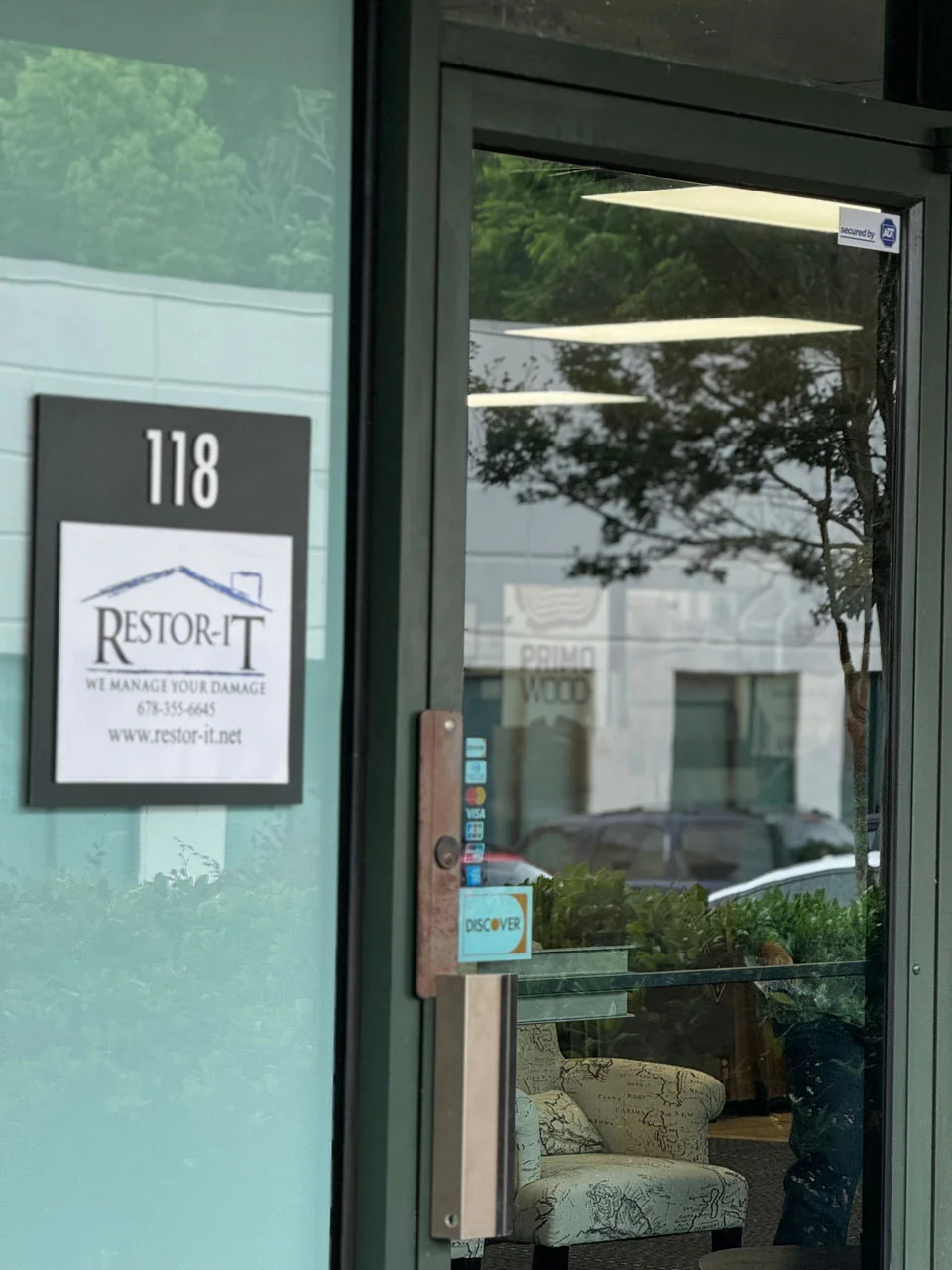 Glass door entrance to a business with the sign "Restor-It" and a door plate numbered "118." A Discover card sticker is visible. The reflection shows outdoor greenery and building exteriors.