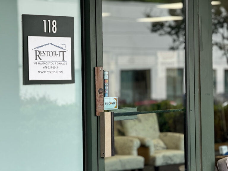 Flooring Installation 9 Glass door with a sign for "Restor-It" and entrance number 118. Inside, there are beige chairs and large windows reflecting outside greenery.