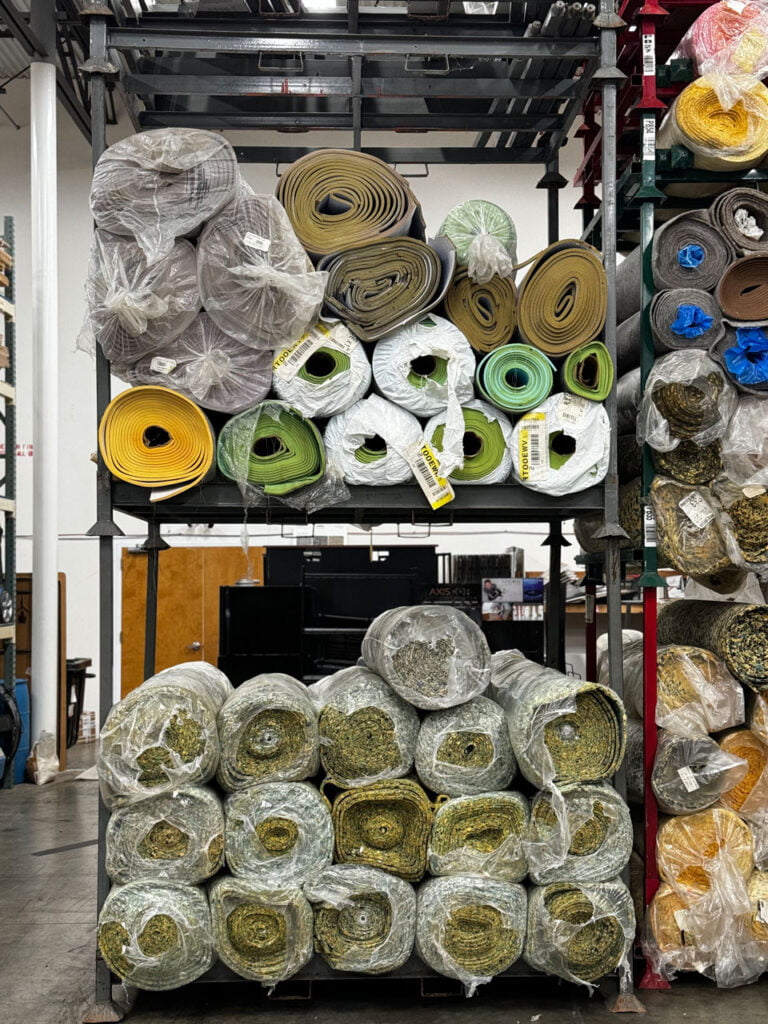 Shelves stacked with rolled-up carpets and mats in various colors and types, wrapped in plastic, inside a warehouse-like environment.