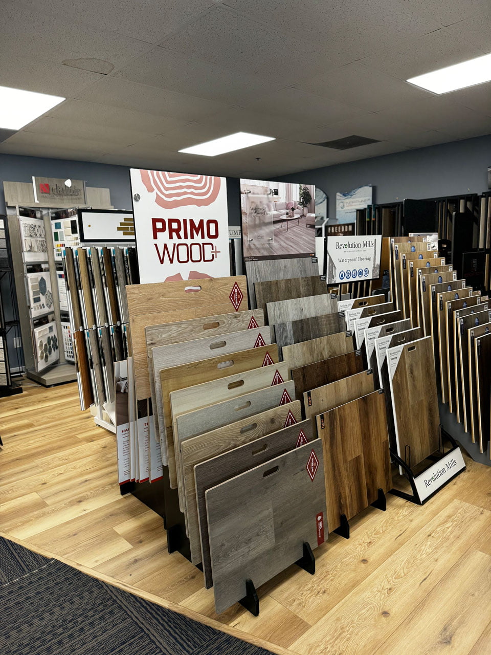 About 6 A store display featuring various wood flooring samples, including a prominent section labeled "Primo Wood+" with different shades and finishes available for customers to view.