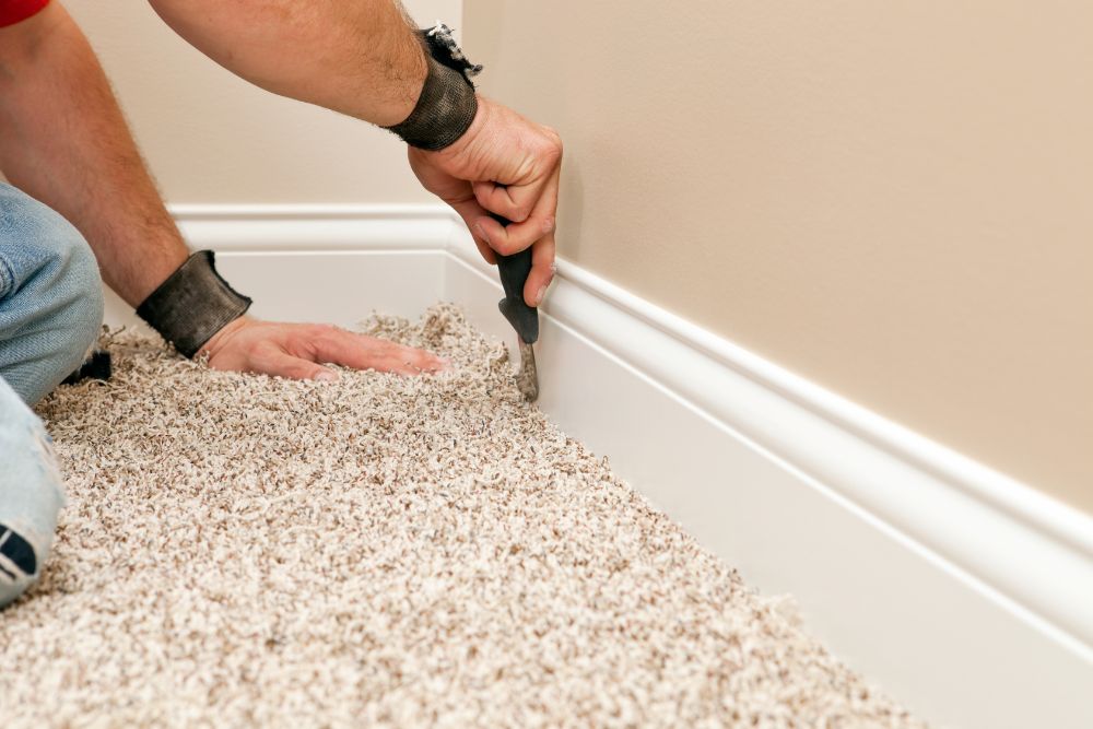 Flooring Installation 10 A person trims the edge of a beige carpet along a white baseboard with a utility knife, showcasing their meticulous attention to detail in flooring installation services.