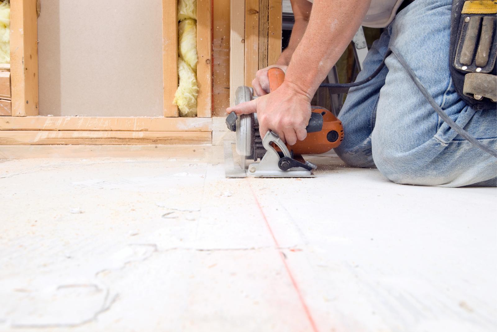 Flooring And Subflooring Repairs 5 A person uses a circular saw to cut a wooden board on the floor, preparing for subfloor repairs. They are kneeling and wearing jeans with a tool belt on their waist.