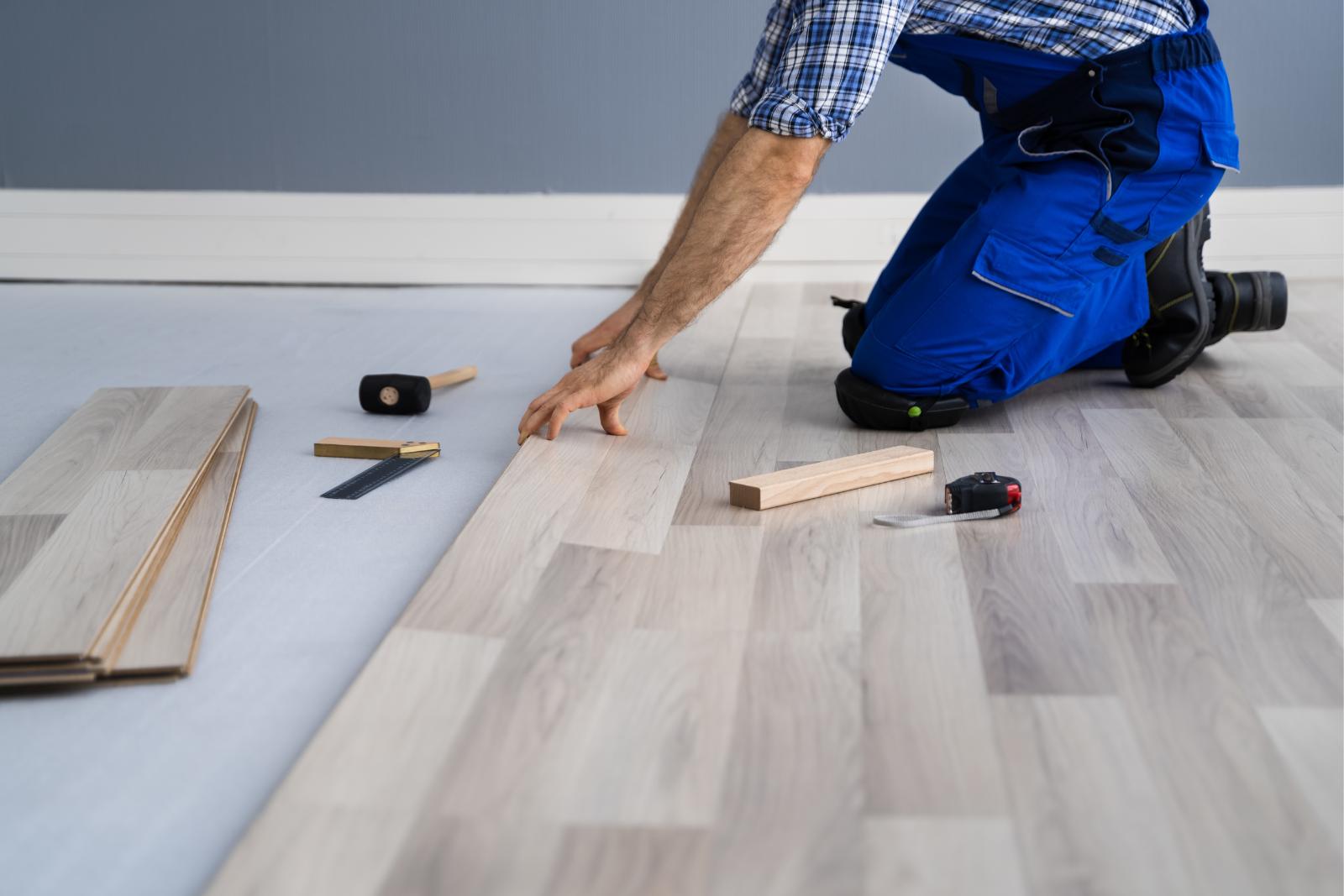 Flooring And Subflooring Repairs 18 A person in blue overalls installs light wood flooring, surrounded by tools and plank pieces, carefully ensuring the subfloor is sturdy and level.