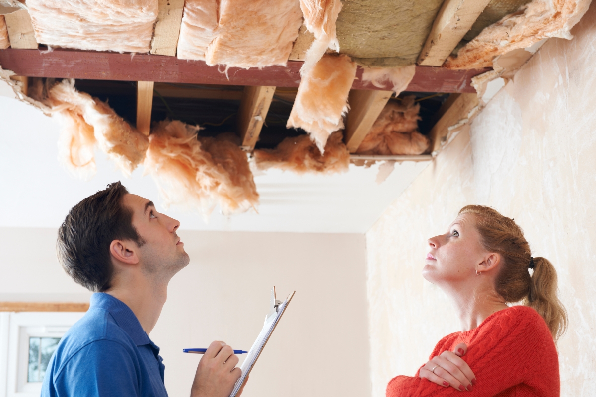 Blog 3 Two people inspecting a ceiling with insulation exposed, one holding a clipboard and pen, assessing the situation for water damage recovery.