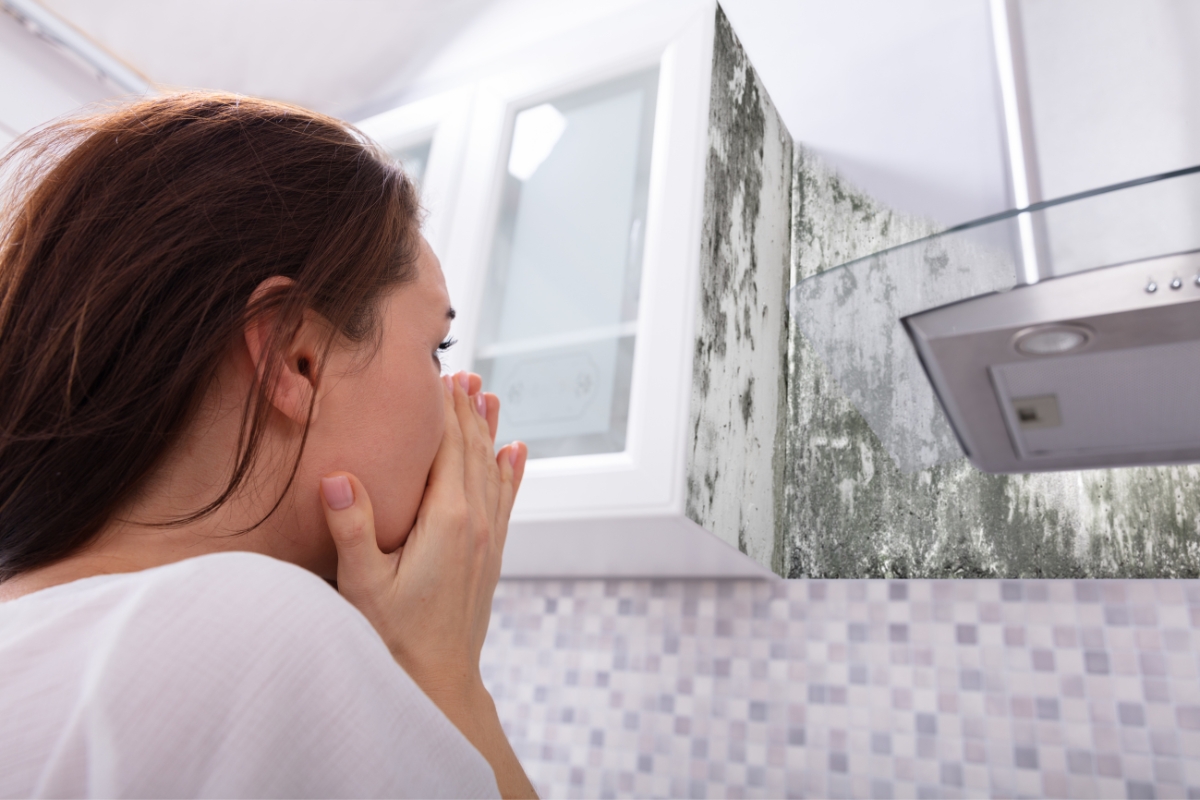 Blog 5 A person covers their mouth, alarmed at the kitchen wall near the oven hood covered in mold, aware of the health risks associated with mold.