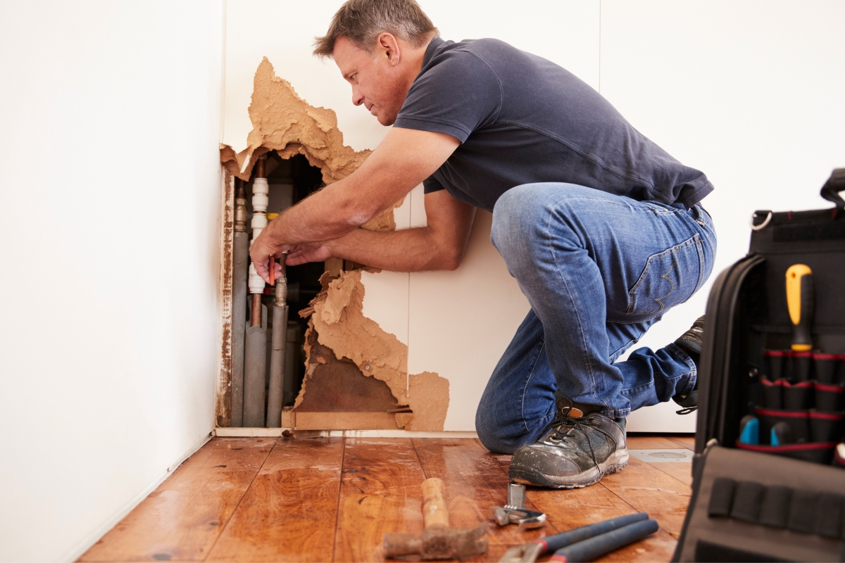 Blog 1 A man kneels while repairing exposed pipes in a damaged wall, demonstrating the precision involved in emergency remediation services. A tool bag and tools are scattered on the wooden floor nearby, ready for swift action.