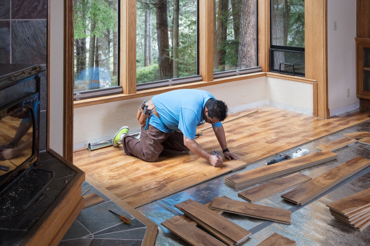 Blog 6 A person expertly installs wooden flooring in a sunlit room with large windows overlooking a serene forest. Their dedication to quality flooring ensures the room's elegance is perfectly highlighted by nature’s backdrop.