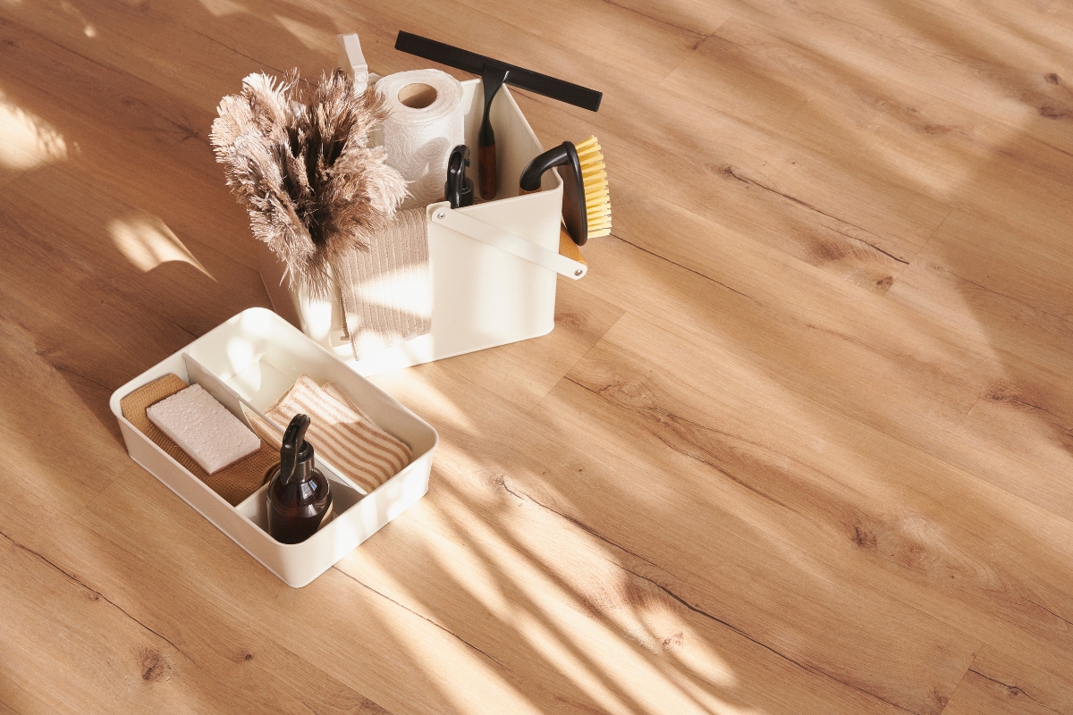 Vinyl Vs Laminate Flooring: Which Is Best For Your Georgia Home? 6 A cozy Georgia home scene: cleaning supplies artfully arranged on a wooden floor, featuring a bottle, brushes, a roll of paper, and cloths in sleek white containers. Pampas grass adds a decorative touch to this laminate flooring vignette.