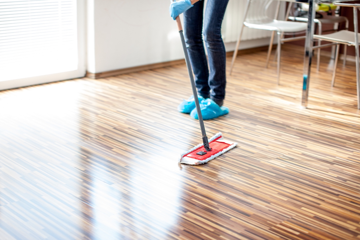 Vinyl Vs Laminate Flooring: Which Is Best For Your Georgia Home? 7 In a charming Georgia home, sunlight streams through the window as a person diligently mops a wooden floor, their blue shoe covers and gloves catching the glow.