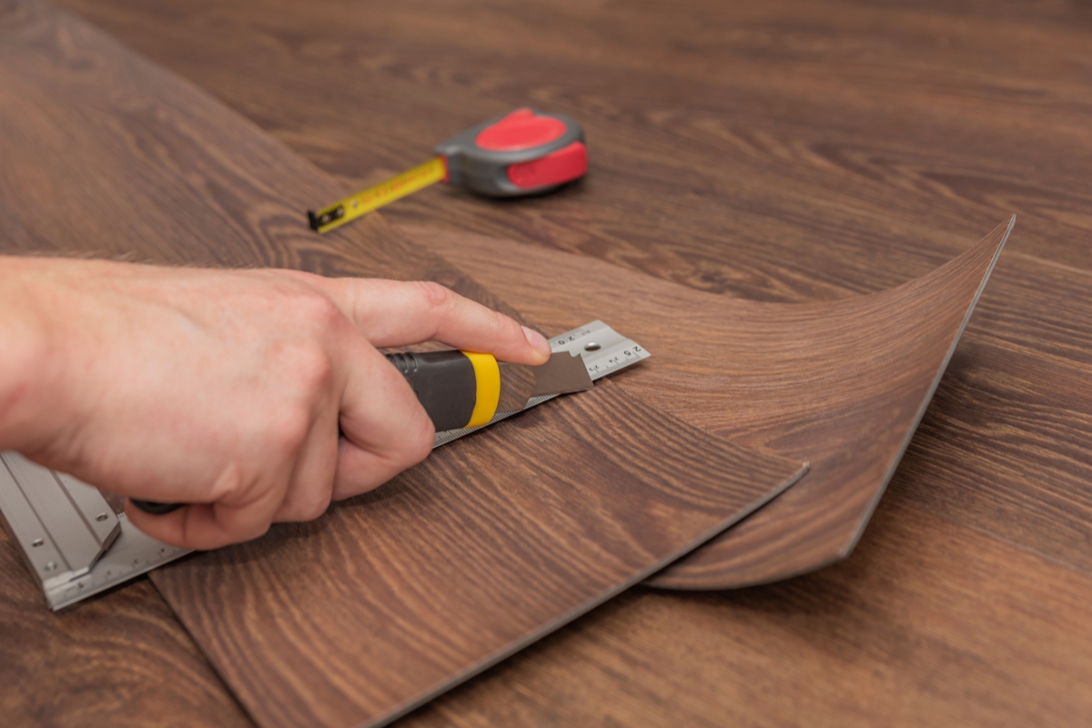 Vinyl Vs Laminate Flooring: Which Is Best For Your Georgia Home? 8 In a Georgia home, a person skillfully uses a utility knife to cut vinyl flooring along a metal ruler, with a tape measure resting on the wooden surface in the background.