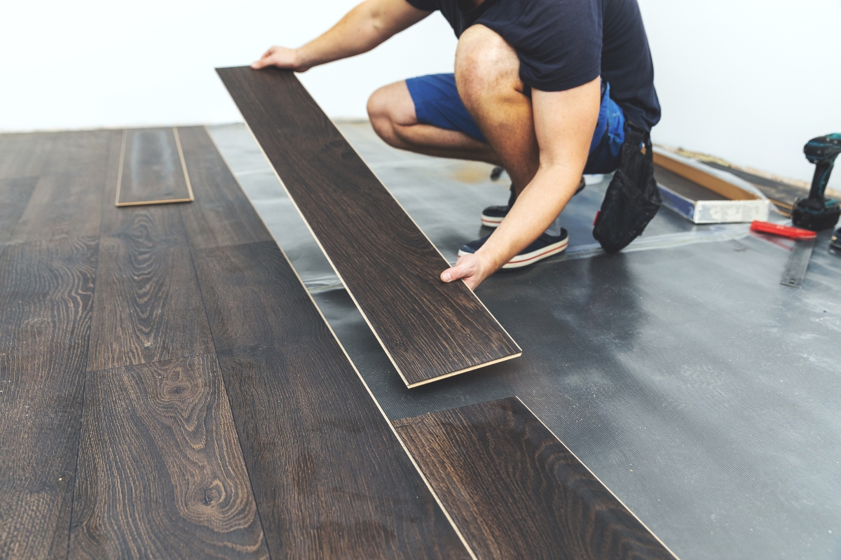 Vinyl Vs Laminate Flooring: Which Is Best For Your Georgia Home? 9 In a cozy Georgia home, someone is installing dark wood laminate flooring, carefully holding a plank in place. Nearby, essential tools lay ready for use.
