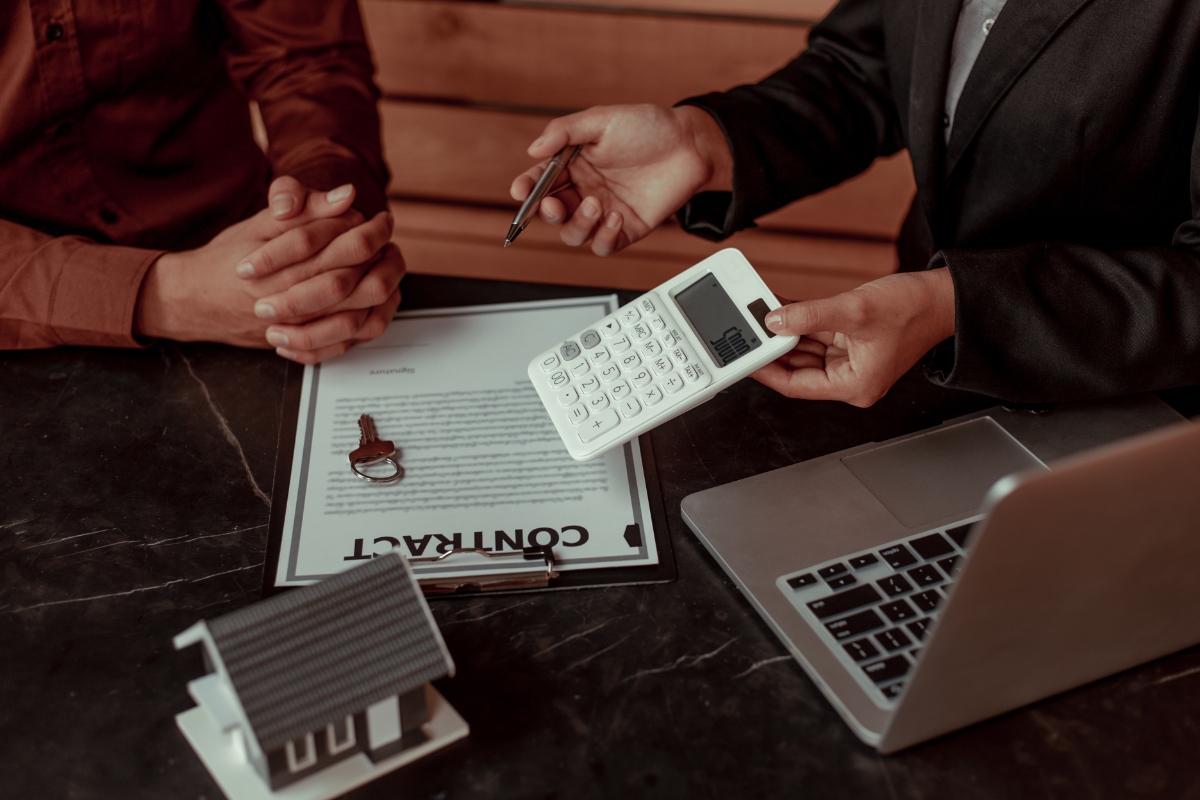 Vinyl Vs Laminate Flooring: Which Is Best For Your Georgia Home? 11 Two people discuss over a contract document, surrounded by a key, small house model, and laptop on the table. The conversation hints at design choices like laminate flooring for their Georgia home, while the calculator crunches numbers in silent agreement.