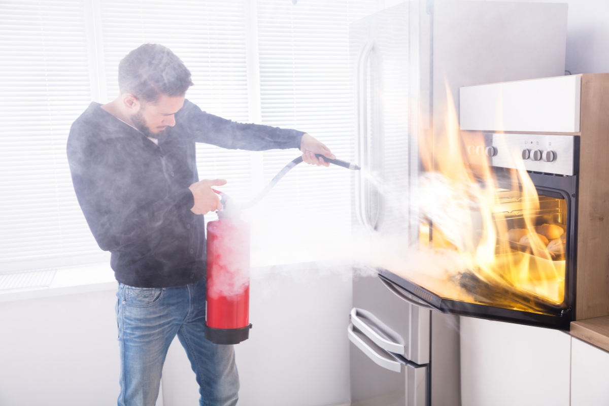 A man uses a fire extinguisher to put out a fire in an oven in a modern kitchen. Smoke and flames are visible, highlighting the urgent need for Fire Damage Restoration.