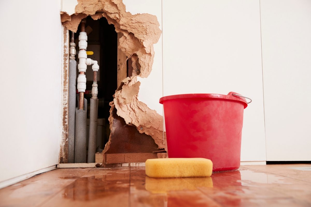 Fire Damage Restoration: Transform Your Home Back To Normal Swiftly 4 A large hole in a wall reveals water pipes, with a red bucket and yellow sponge on a wet floor in front of it—ideal for Fire Damage Restoration professionals to assess and repair both visible and hidden damage.