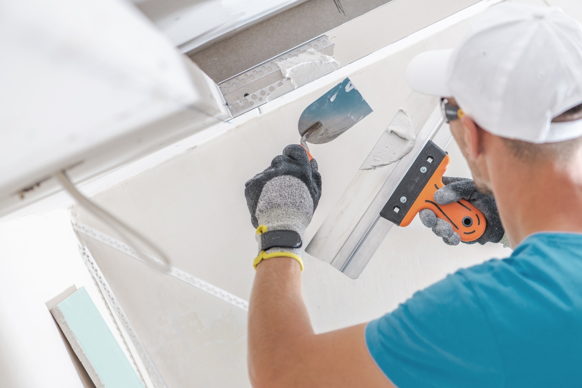 A person in gloves and a cap uses a drywall taping knife to expertly spread joint compound on a ceiling corner, showcasing Professional Drywall Repair skills during construction or renovation work.