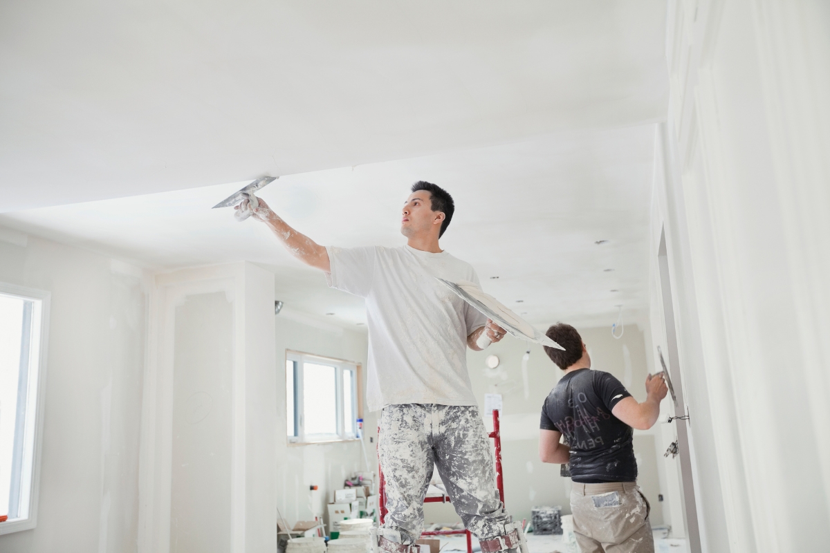 Professional Drywall Repair For A Smooth And Flawless Home Interior 2 Two people in paint-stained clothes apply plaster or paint to the ceiling and wall of a bright, unfinished room, showcasing their expertise in professional drywall repair.