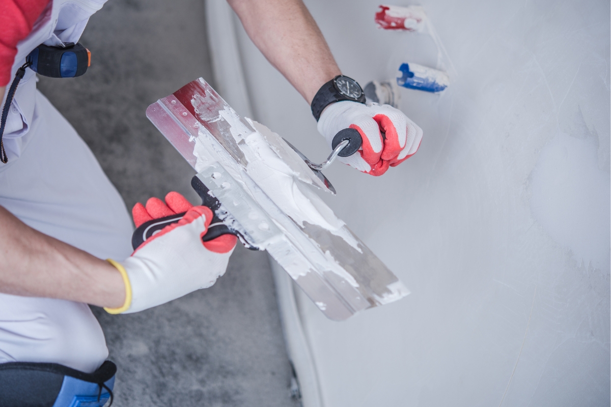 Professional Drywall Repair For A Smooth And Flawless Home Interior 3 A person wearing gloves spreads plaster onto a large metal putty knife using a smaller tool, showcasing the careful techniques used in professional drywall repair.