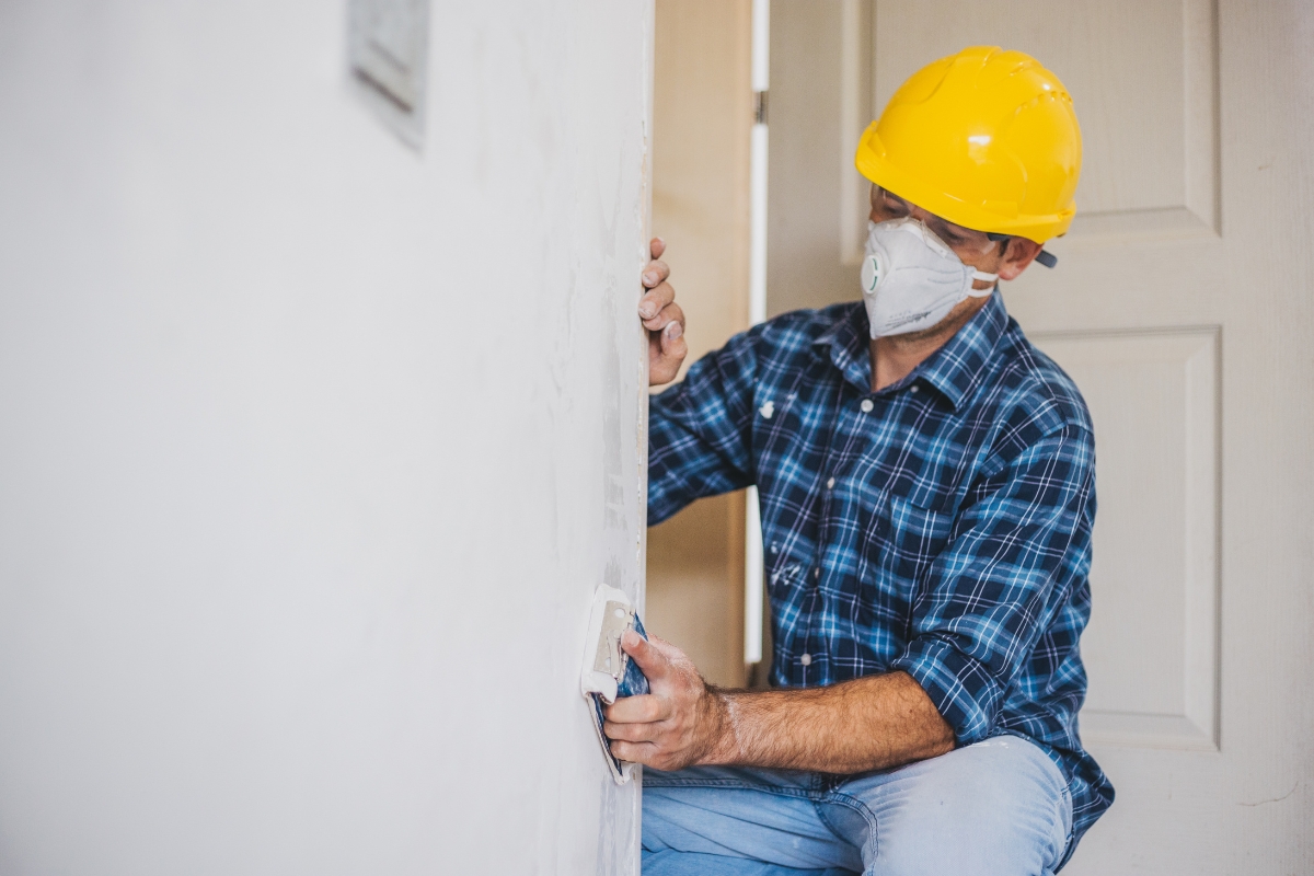 Professional Drywall Repair For A Smooth And Flawless Home Interior 4 A person wearing a hard hat, face mask, and plaid shirt sands a white wall in a room under construction, showcasing professional drywall repair skills.