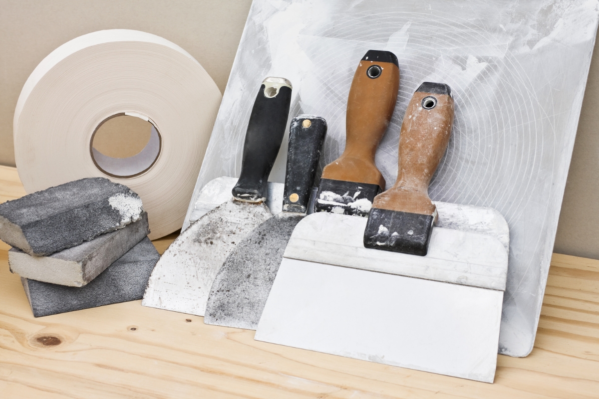 Professional Drywall Repair For A Smooth And Flawless Home Interior 7 A selection of professional drywall repair tools—taping knives, putty knives, sanding sponges, a roll of joint tape, and a mud pan—is neatly arranged on a wooden surface.