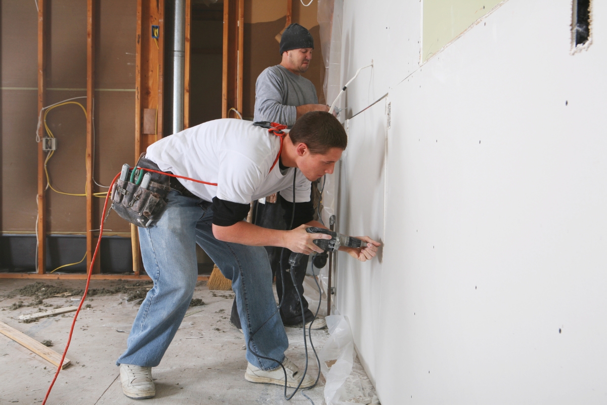 Professional Drywall Repair For A Smooth And Flawless Home Interior 8 Two workers perform professional drywall repair; one uses a power tool to cut out a section while the other stands nearby observing in a partially finished room.