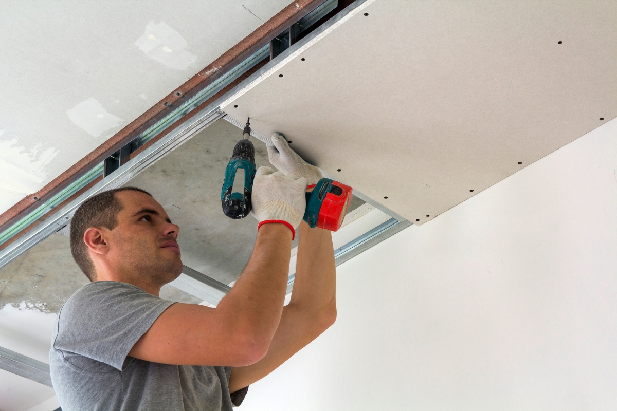 Professional Drywall Repair For A Smooth And Flawless Home Interior 1 A man installs a sheet of drywall on a ceiling using a power drill while wearing white work gloves, demonstrating expert Professional Drywall Repair skills.