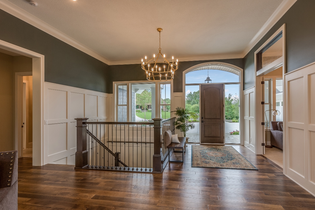 Entryway of a house with vinyl plank flooring, a staircase, chandelier, green walls with white wainscoting, open front door, and windows showing greenery outside.