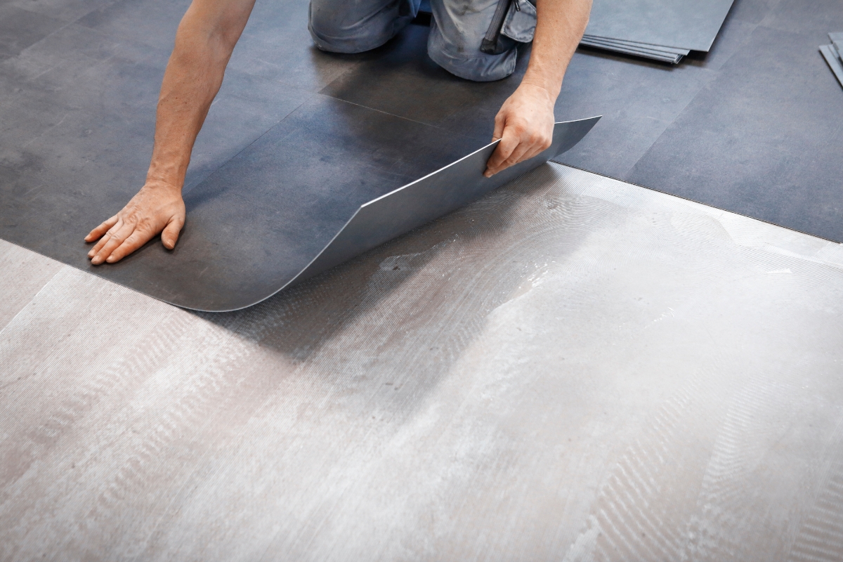 Luxury Vinyl Plank Flooring: Transform Modern Homes With Style And Durability 2 A person installs Vinyl Plank Flooring, carefully placing a dark tile over a light subfloor. Only their hands and knees are visible.