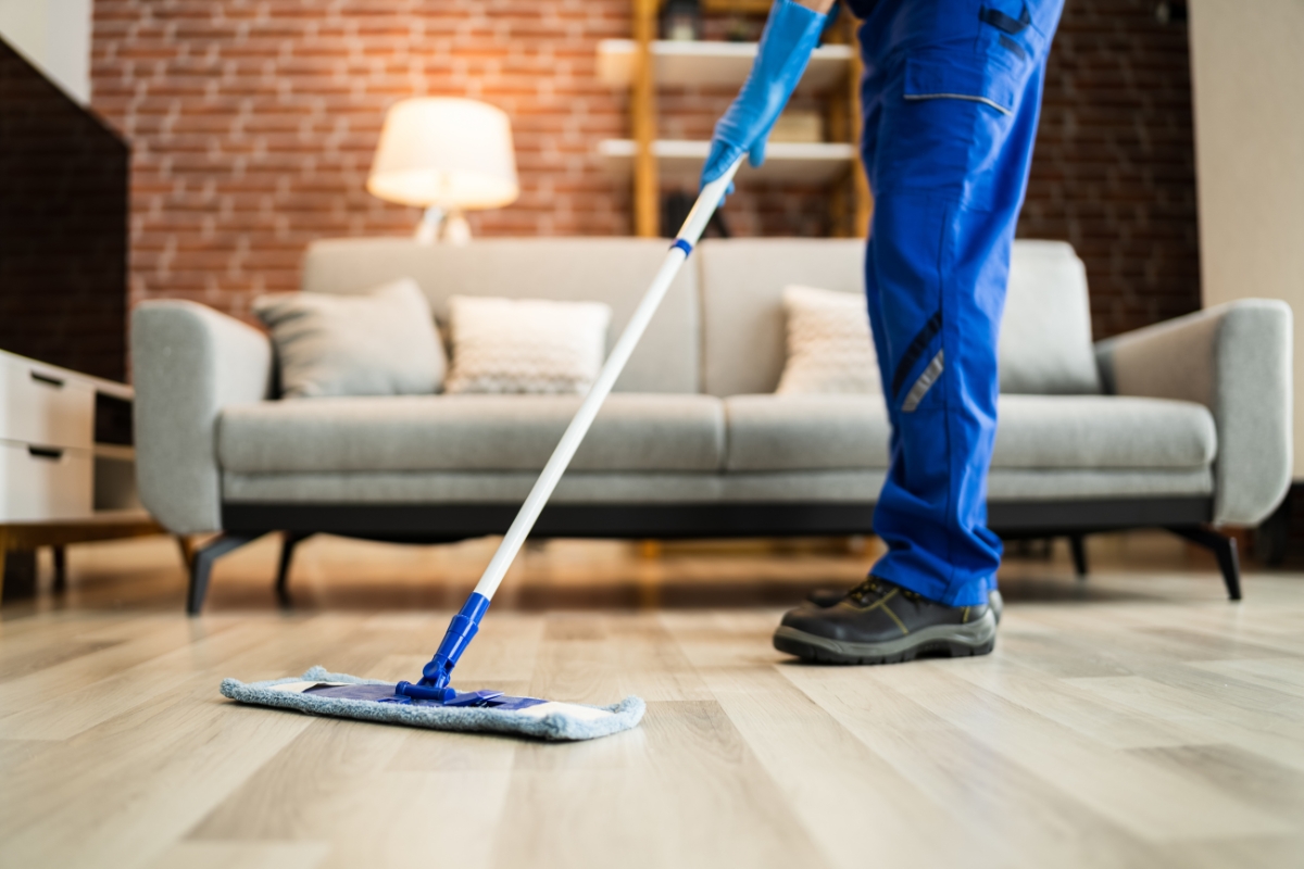 Luxury Vinyl Plank Flooring: Transform Modern Homes With Style And Durability 4 Person in blue work clothes mopping a vinyl plank flooring in a living room with a gray sofa, brick wall, and lit floor lamp in the background.