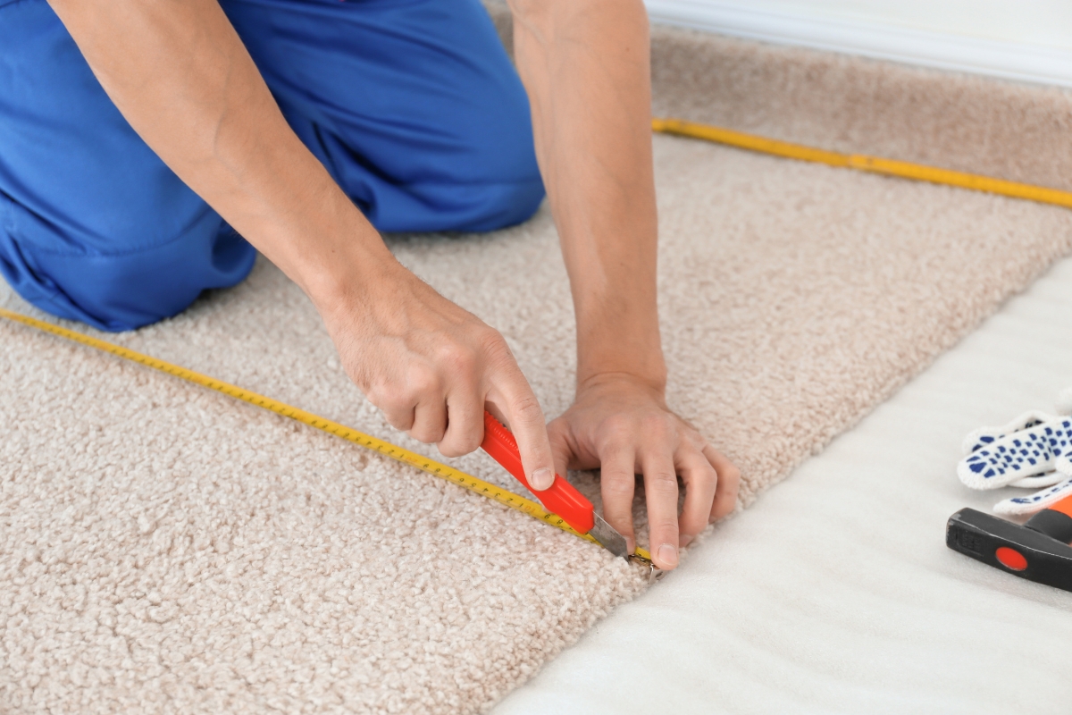 Carpet Replacement: Top Signs It’s Time For A New Carpet In Your Home 6 Person using a utility knife to cut beige carpet along a measuring tape, with flooring tools nearby—a precise step in the carpet replacement process.