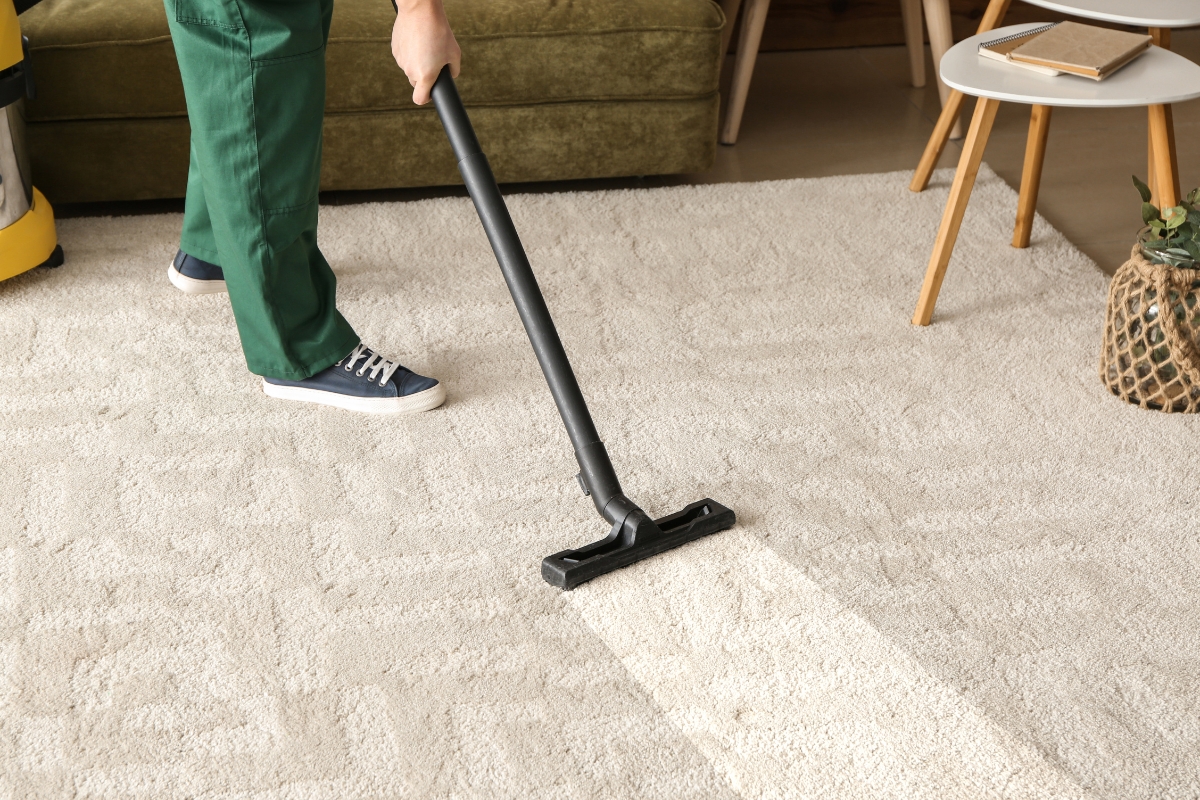 Carpet Replacement: Top Signs It’s Time For A New Carpet In Your Home 7 A person wearing green pants and blue sneakers is vacuuming a light-colored carpet in a living room near a brown couch and small table, perhaps preparing the space for a potential carpet replacement.