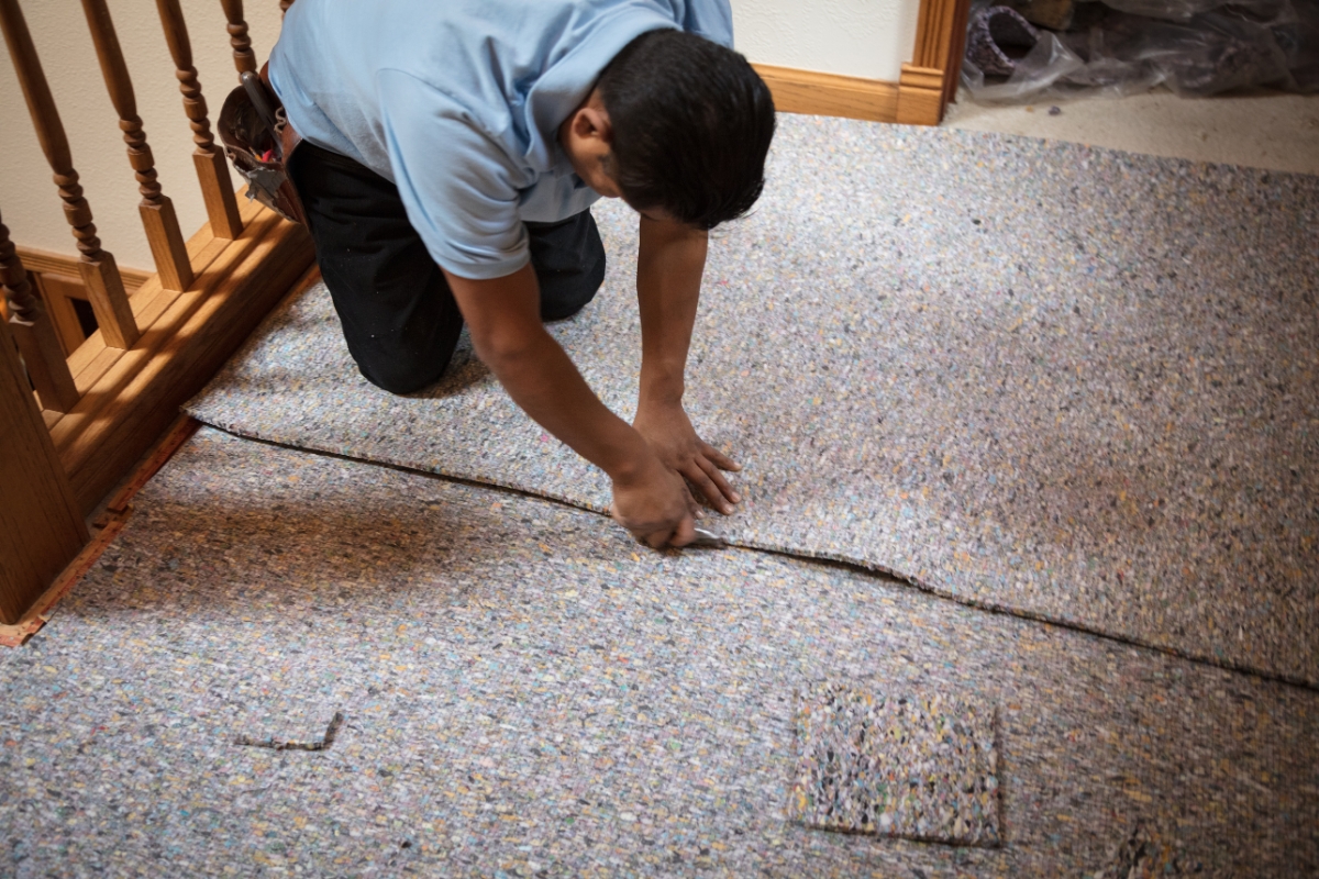 Carpet Replacement: Top Signs It’s Time For A New Carpet In Your Home 8 A person kneels on the floor, carefully aligning and installing carpet padding near a wooden railing as part of a carpet replacement inside a house.
