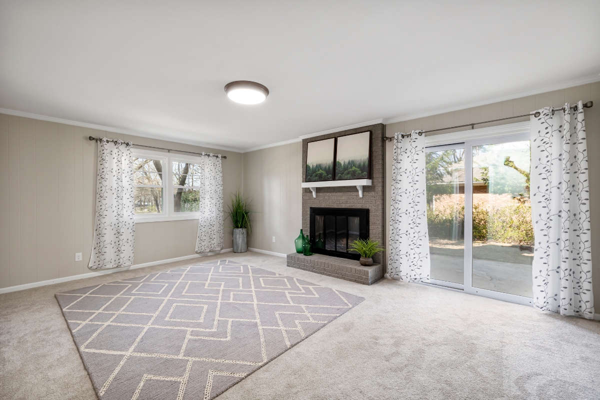Bright living room with neutral walls, patterned curtains, large windows, sliding glass door, geometric area rug over recent carpet replacement, fireplace, and minimal decor.