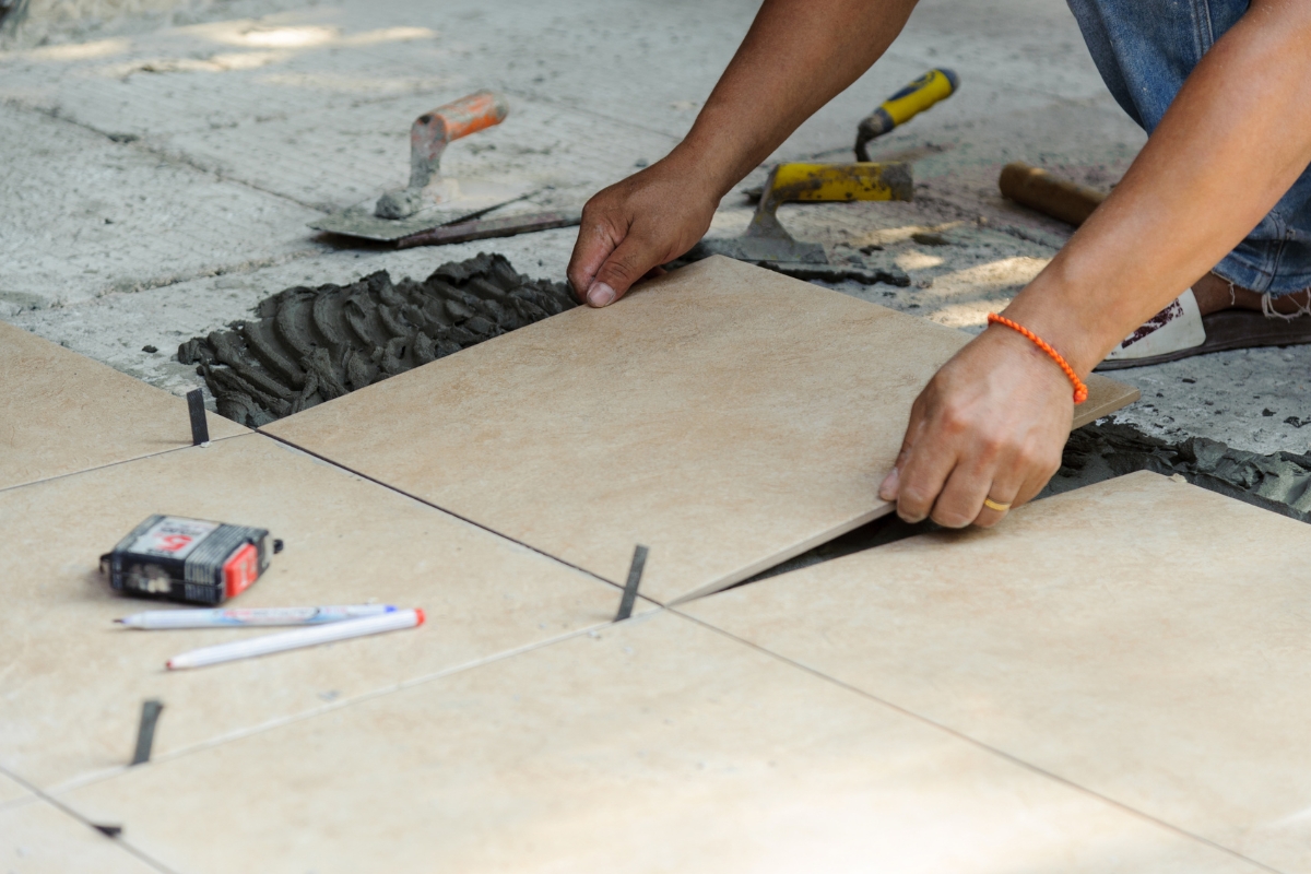Tile Flooring Installation: Why It’s A Great Choice For Atlanta Homes 17 A person is laying beige ceramic floor tiles on mortar, using spacers for alignment during tile flooring installation. Nearby are tools including a tape measure, trowel, pencil, and hammer.