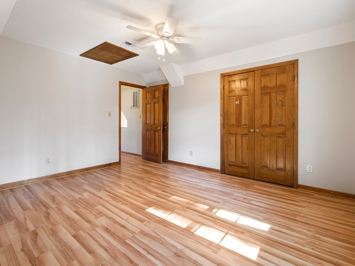Best Flooring For High-Traffic Areas: A Homeowner’s Guide To Durability And Style 11 Empty room with light wood flooring—ideal as the best flooring for high-traffic areas—beige walls, a ceiling fan, two wooden doors, and a ceiling attic access panel. Sunlight streams in through an unseen window.