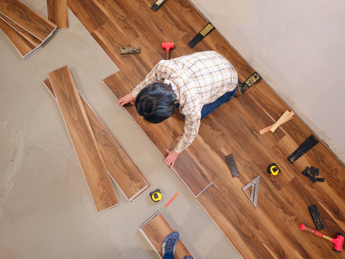 Best Flooring For High-Traffic Areas: A Homeowner’s Guide To Durability And Style 14 A person installs wood laminate flooring—one of the best flooring options for high-traffic areas—over a concrete floor, surrounded by tools like a tape measure, hammer, and squares.