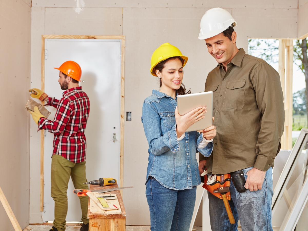 Atlanta Home Renovation Experts: What To Expect From Your Remodel 3 Two construction workers in hard hats review a tablet together while another installs drywall in the background, showcasing teamwork on an Atlanta Home Renovation project.