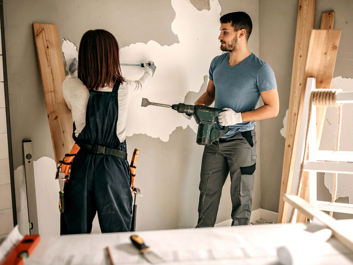Atlanta Home Renovation Experts: What To Expect From Your Remodel 5 Two people renovating a room; one measures the wall while the other holds a power drill. Tools and construction materials are visible, creating the perfect scene for an Atlanta Home Renovation project in progress.