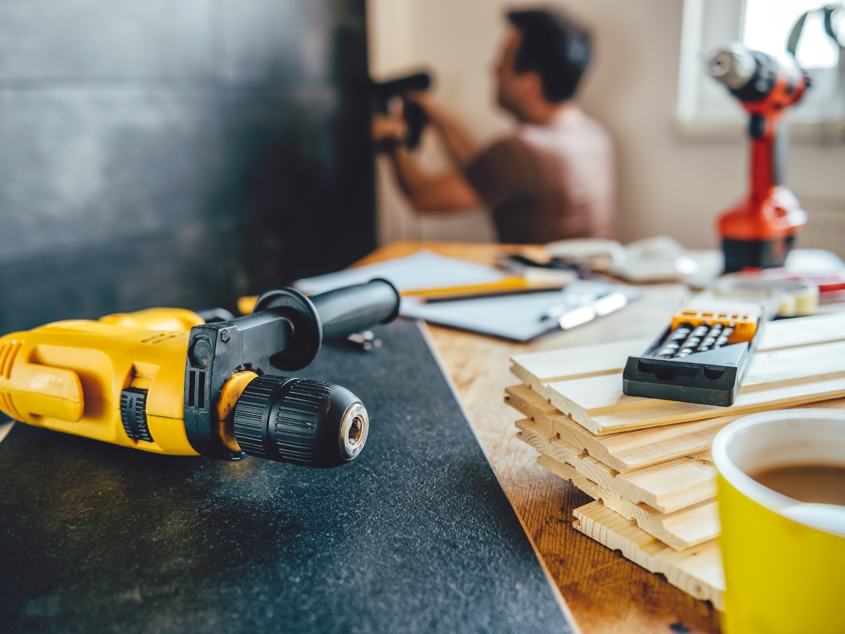 Atlanta Home Renovation Experts: What To Expect From Your Remodel 6 A yellow power drill lies on a workbench with wood planks, a calculator, and tools—typical sights during an Atlanta Home Renovation. In the background, a person uses a drill near the wall.