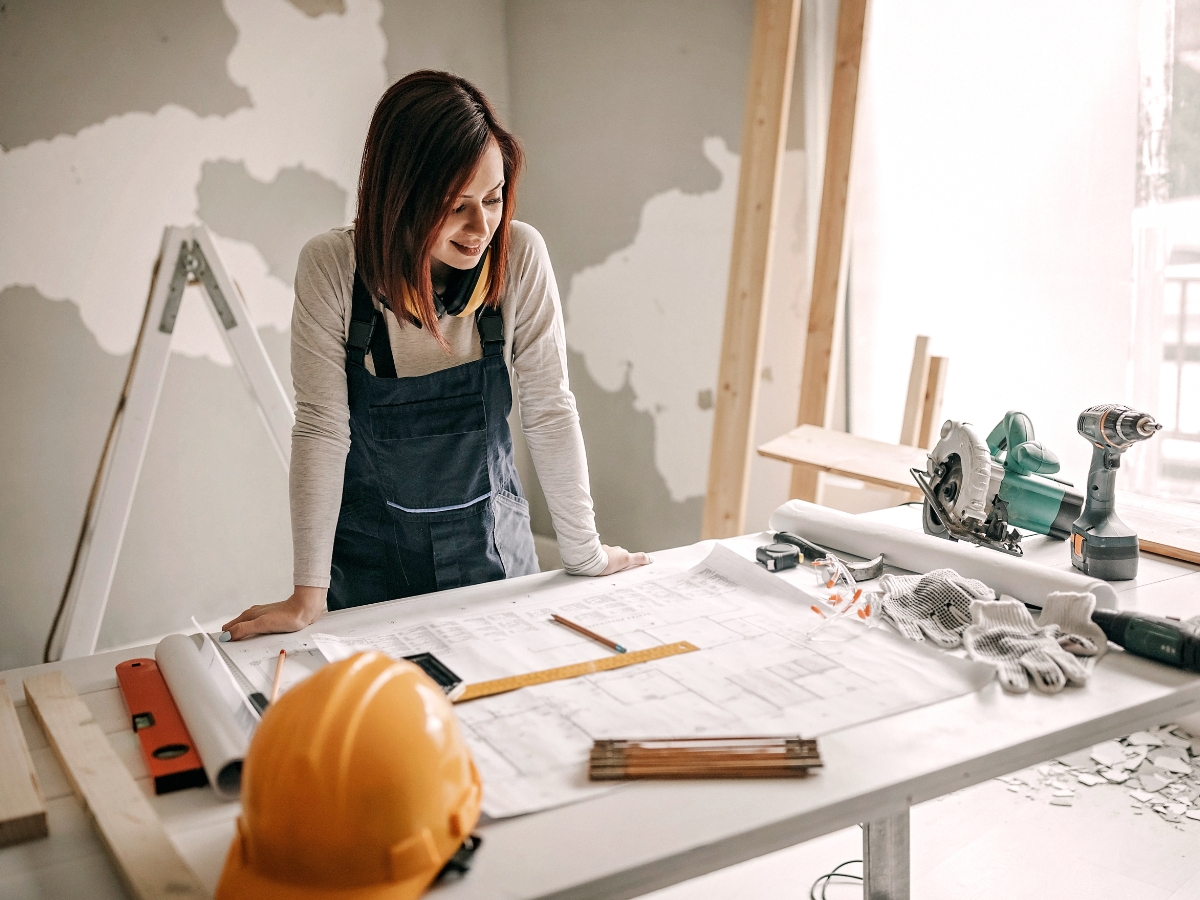 A woman in overalls stands at a worktable with construction plans, tools, and a hard hat in a room under renovation, ready to tackle her latest Atlanta Home Renovation project.