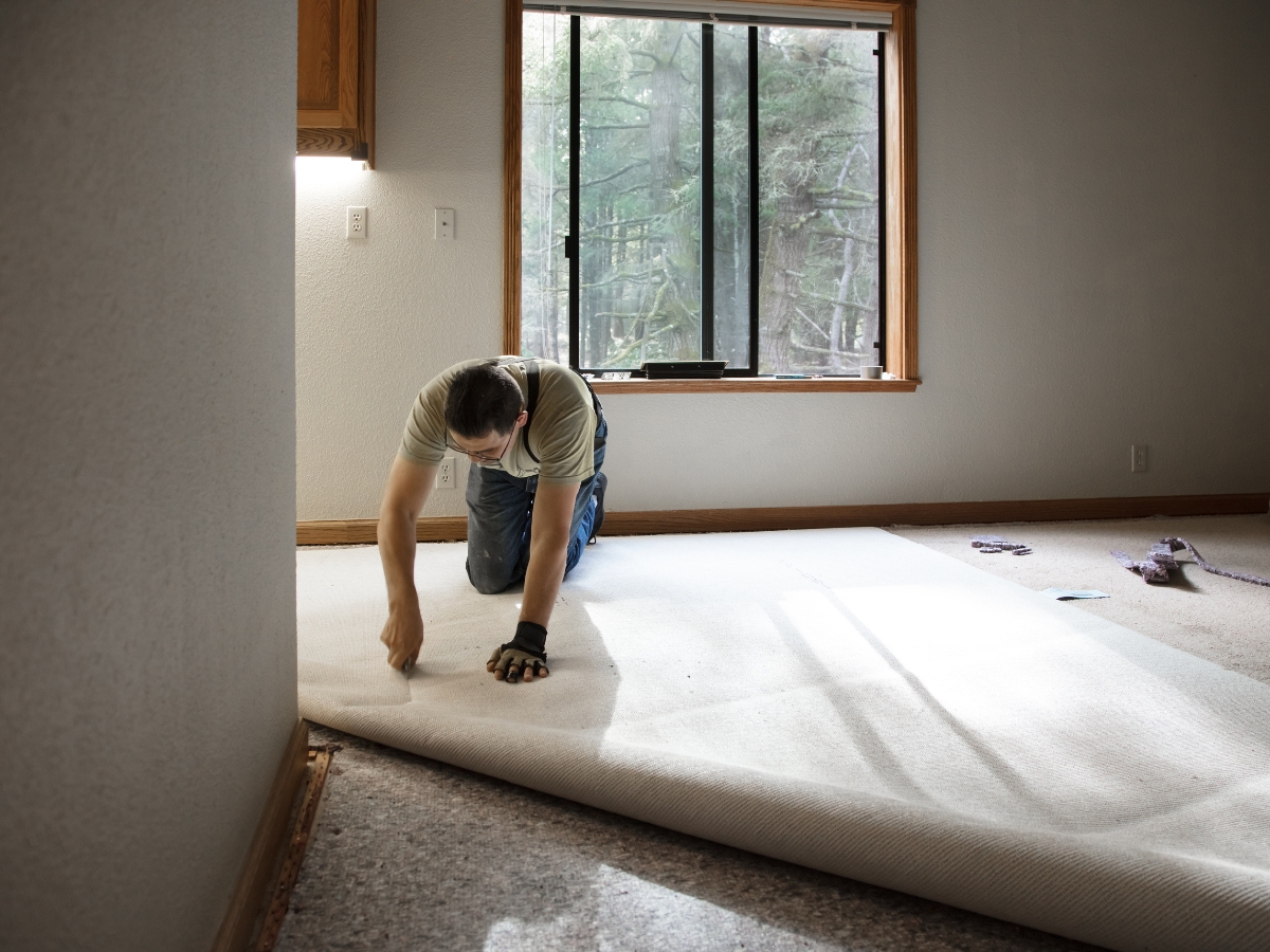 How Professional Carpet Stretching Can Extend The Life Of Your Floors 2 A person kneels on the floor, performing professional carpet stretching while installing new carpet in a room filled with natural light from a large window.
