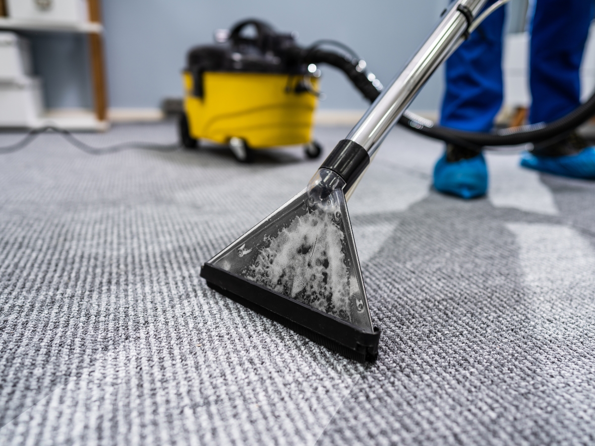 How Professional Carpet Stretching Can Extend The Life Of Your Floors 4 A person uses a carpet cleaning machine with soap and water to clean a gray carpet, while a yellow vacuum sits in the background—perfect maintenance after professional carpet stretching.