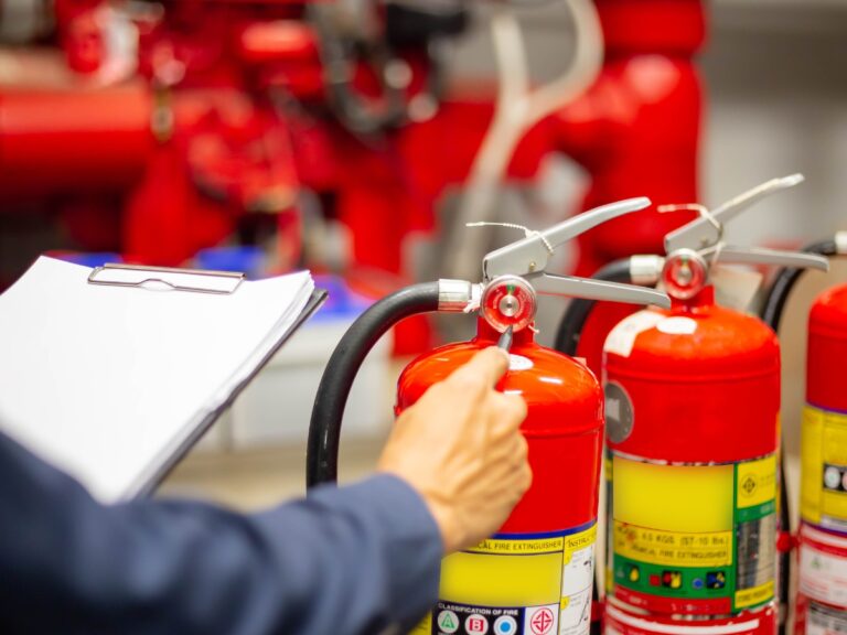 A person inspects red fire extinguishers, holding a clipboard and turning a nozzle, demonstrating essential Fire Prevention Tips with industrial equipment visible in the background.