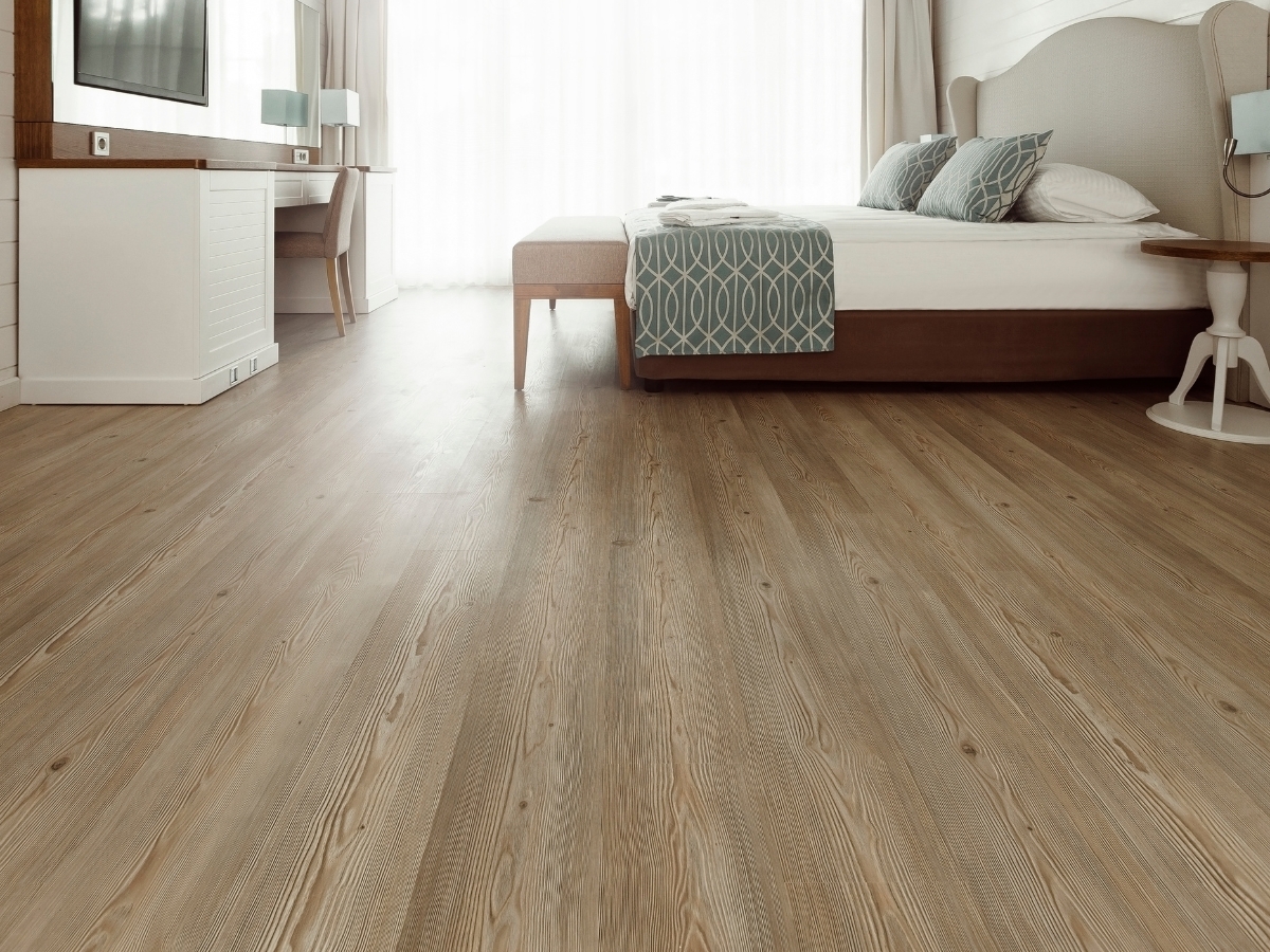 Hardwood Vs. Engineered Wood Flooring: Which One Is Right For You? 2 A modern bedroom with light wood flooring—perfect for those weighing hardwood vs. engineered wood flooring—a bed with patterned bedding, a wall-mounted TV, a desk, and large windows with sheer curtains.