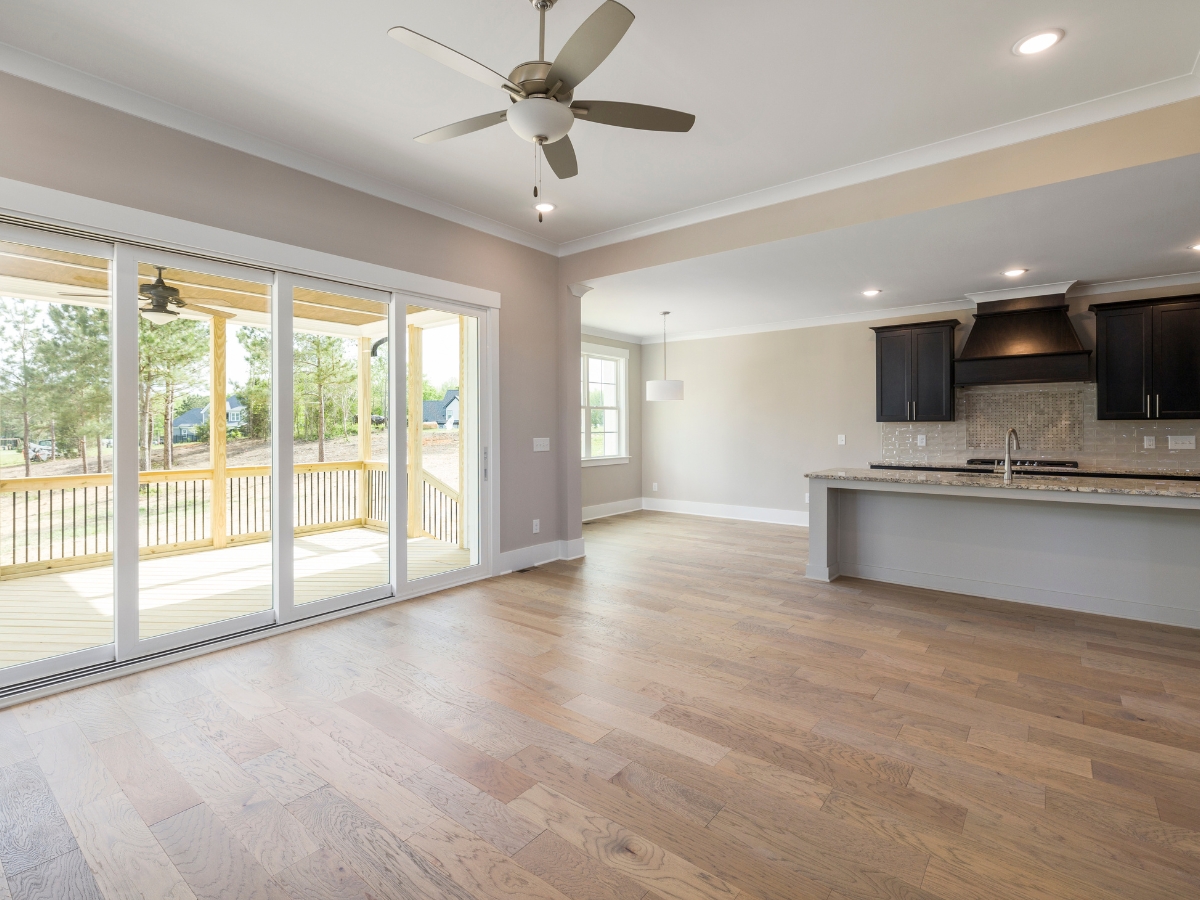 Hardwood Vs. Engineered Wood Flooring: Which One Is Right For You? 3 Spacious empty living area with light wood flooring—perfect for those considering hardwood vs. engineered wood flooring—ceiling fan, large sliding glass doors, and an open view of the kitchen with dark cabinets and a breakfast bar.