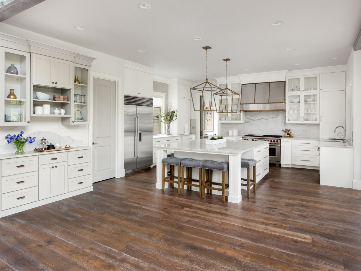 Hardwood Vs. Engineered Wood Flooring: Which One Is Right For You? 4 Modern kitchen with white cabinets, stainless steel appliances, a large island with stools, pendant lights, and striking dark wood flooring—perfect for showcasing the elegance of Hardwood vs. Engineered Wood Flooring options.