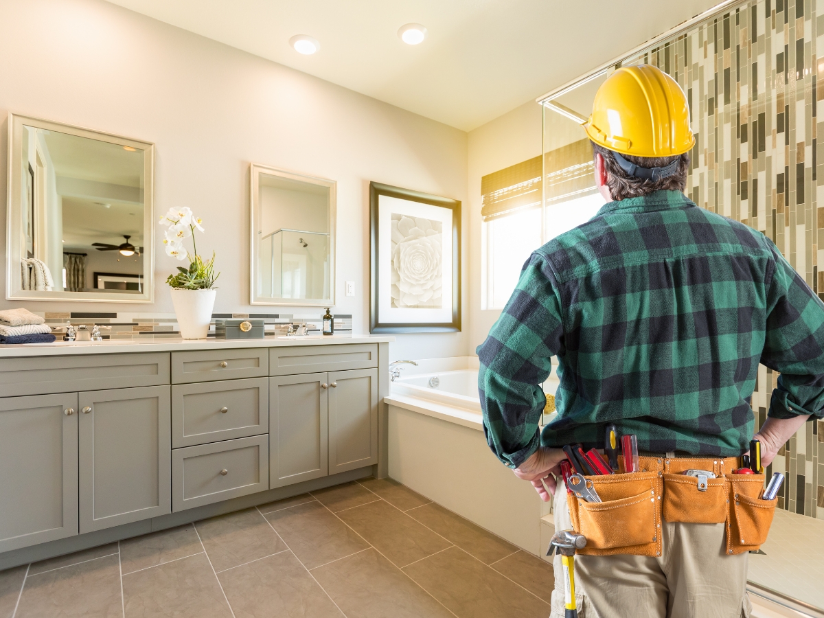 A construction worker wearing a hard hat and tool belt stands in a modern bathroom, evaluating the cabinets, bathtub, and large mirrors under recessed lighting—a scene inspired by Bathroom Remodeling Trends for 2025.