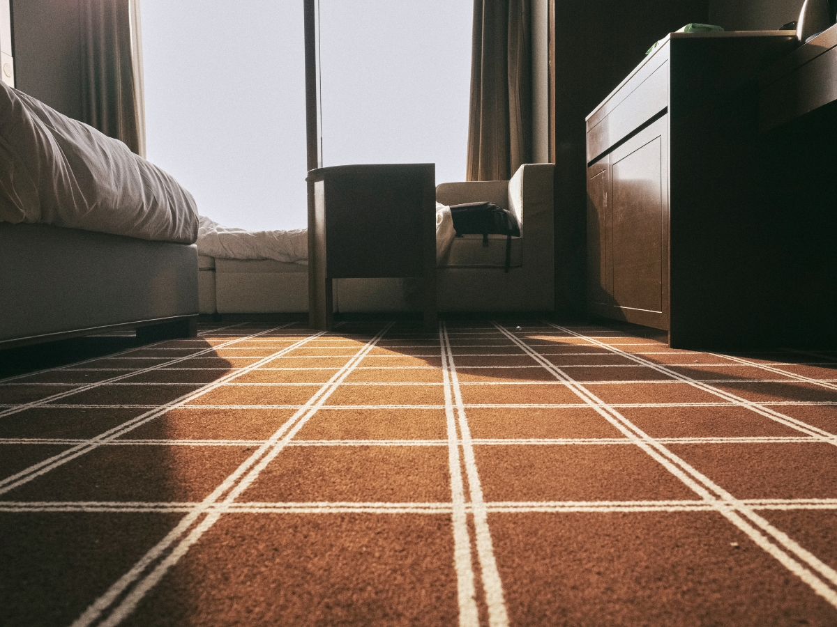 How To Choose The Best Carpet For Home Interior Design 2 A low-angle view of a hotel room with a bed, desk, chair, and a window letting in natural light onto the best carpet for home interior design, showcasing its stylish, patterned look.