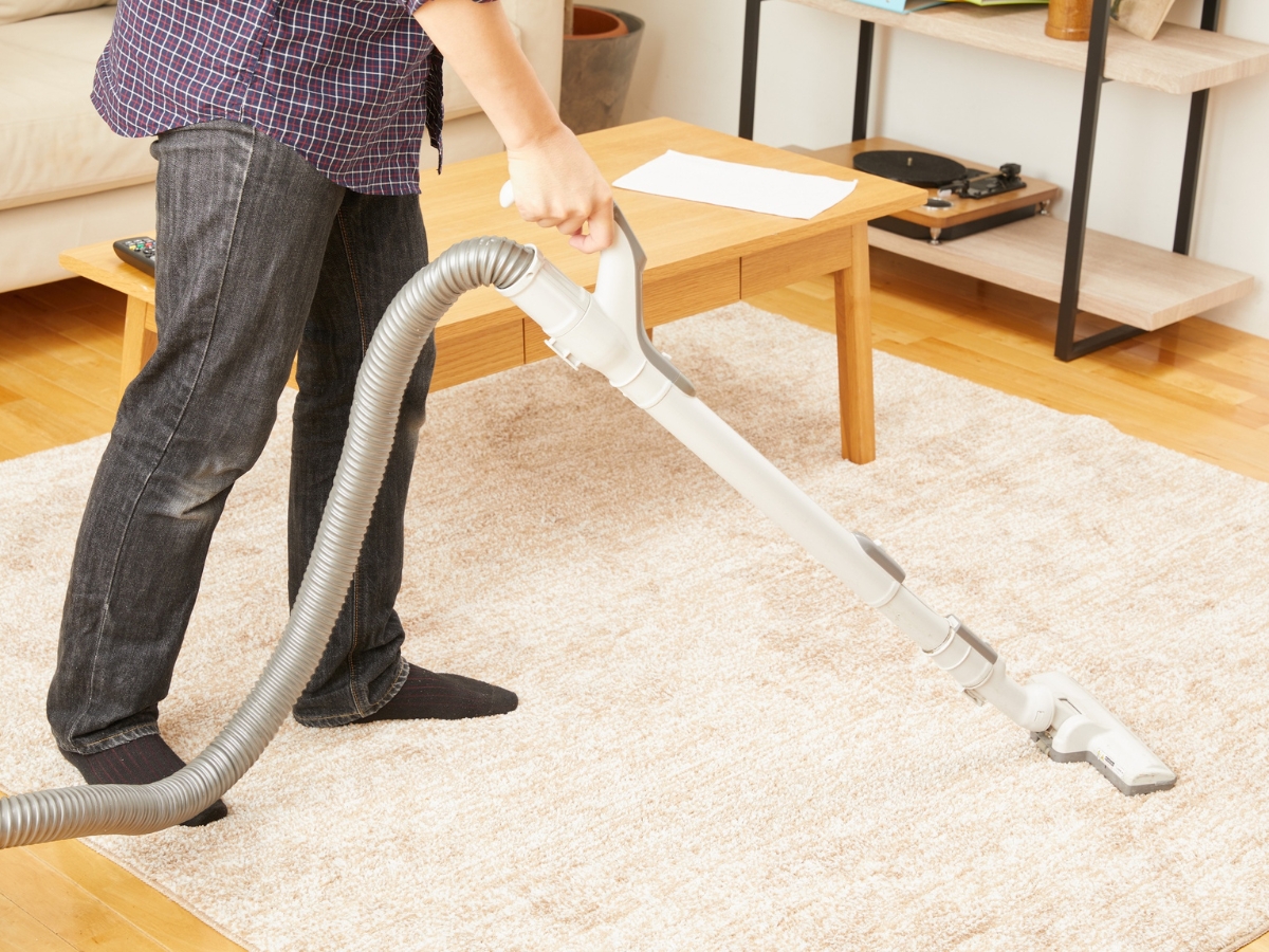 How To Choose The Best Carpet For Home Interior Design 4 A person vacuuming a light-colored rug, considered one of the best carpets for home interior design, in a living room with a coffee table and shelves in the background.