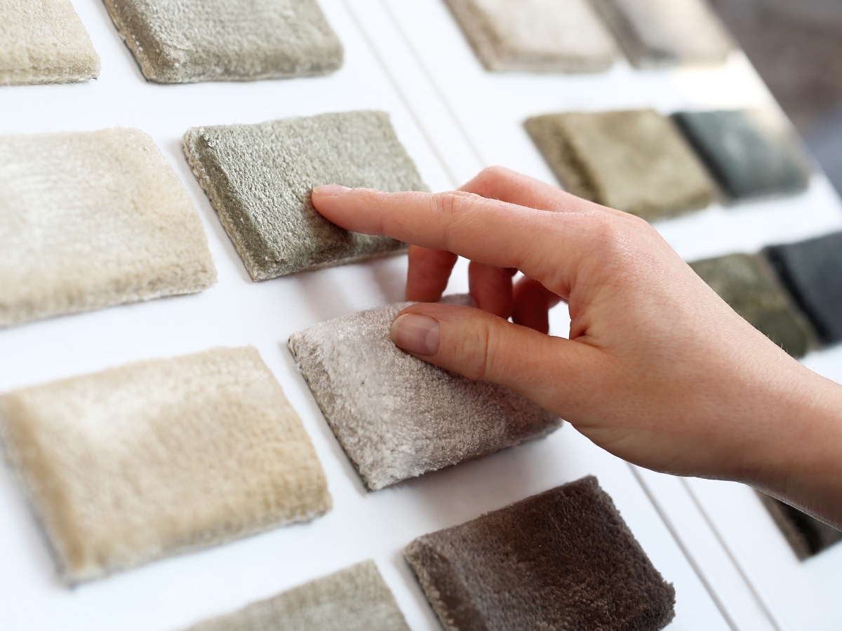 How To Choose The Best Carpet For Home Interior Design 6 A hand touches a sample of carpet swatches arranged in rows on a display board, helping to choose the best carpet for home interior design with various neutral colors and textures.