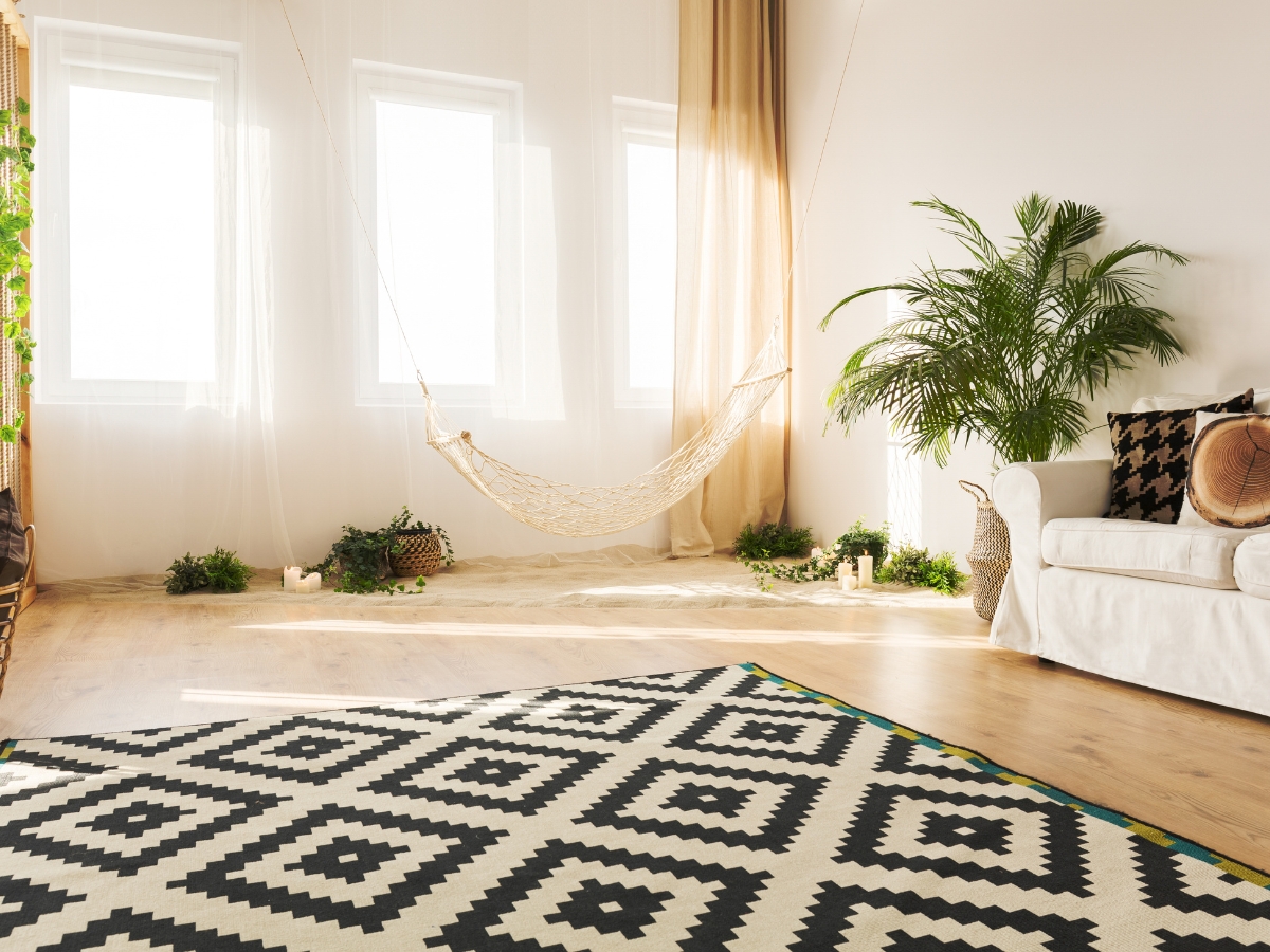 How To Choose The Best Carpet For Home Interior Design 1 A bright living room with large windows, a hammock, potted plants, and a white sofa features the best carpet for home interior design—a striking black-and-white geometric patterned rug.